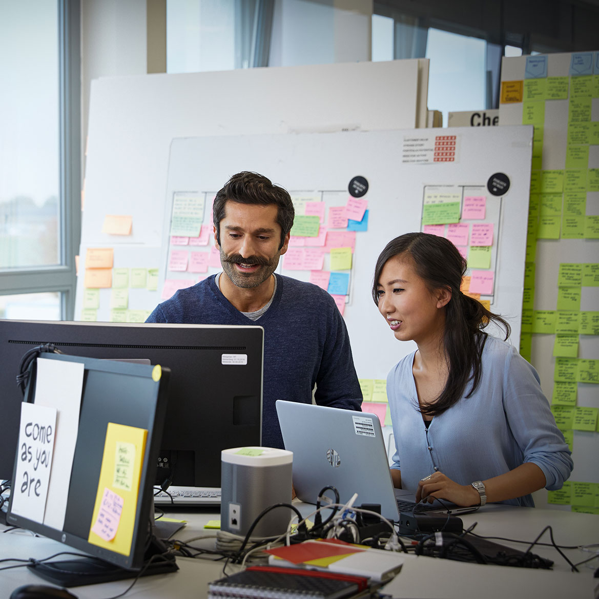 The image shows two colleagues having a meeting at their desk.