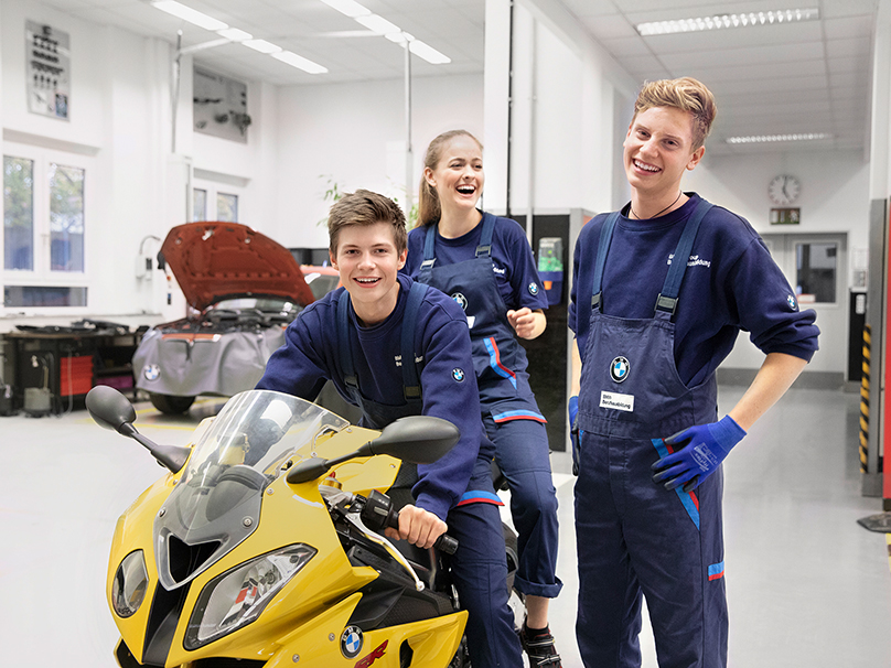Das Bild zeigt drei lachende Auszubildende in einer BMW Werkstatt.