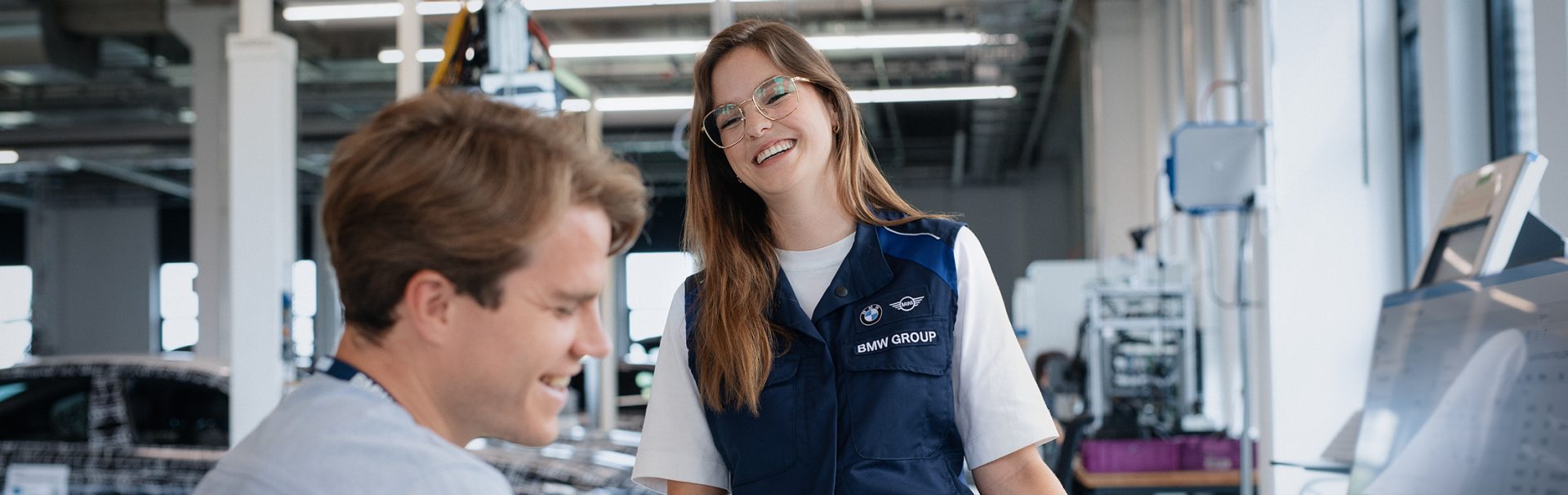 Zwei Studierende arbeiten und lachen in einer Werksumgebung.