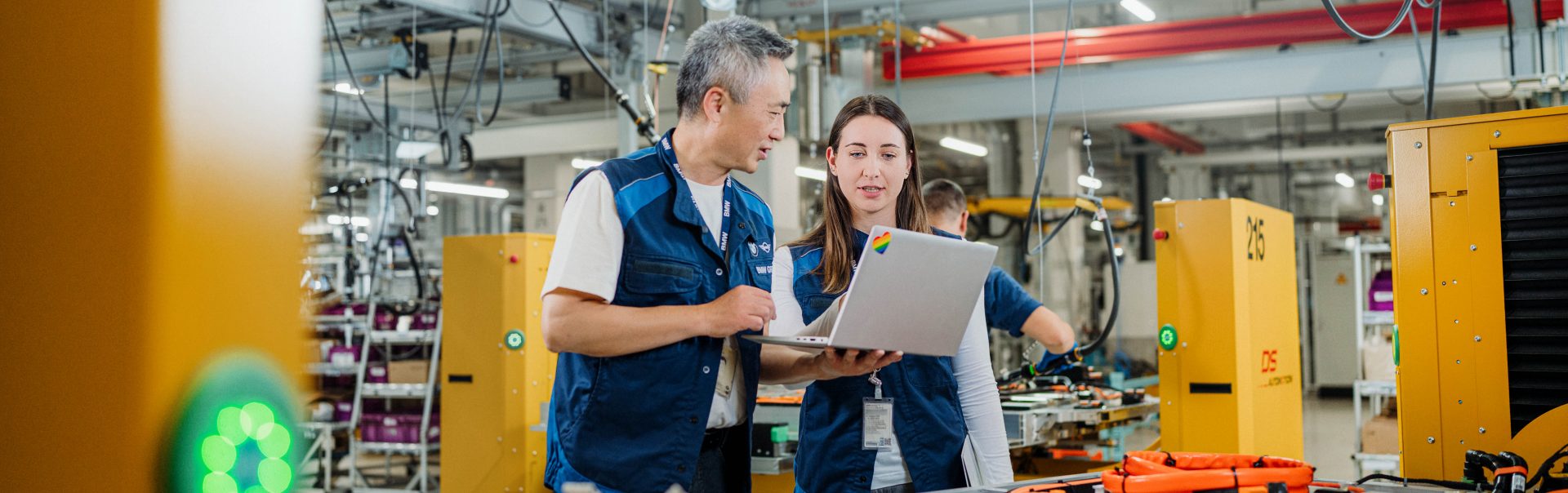 Two BMW employees are working on a laptop in a factory setting.
