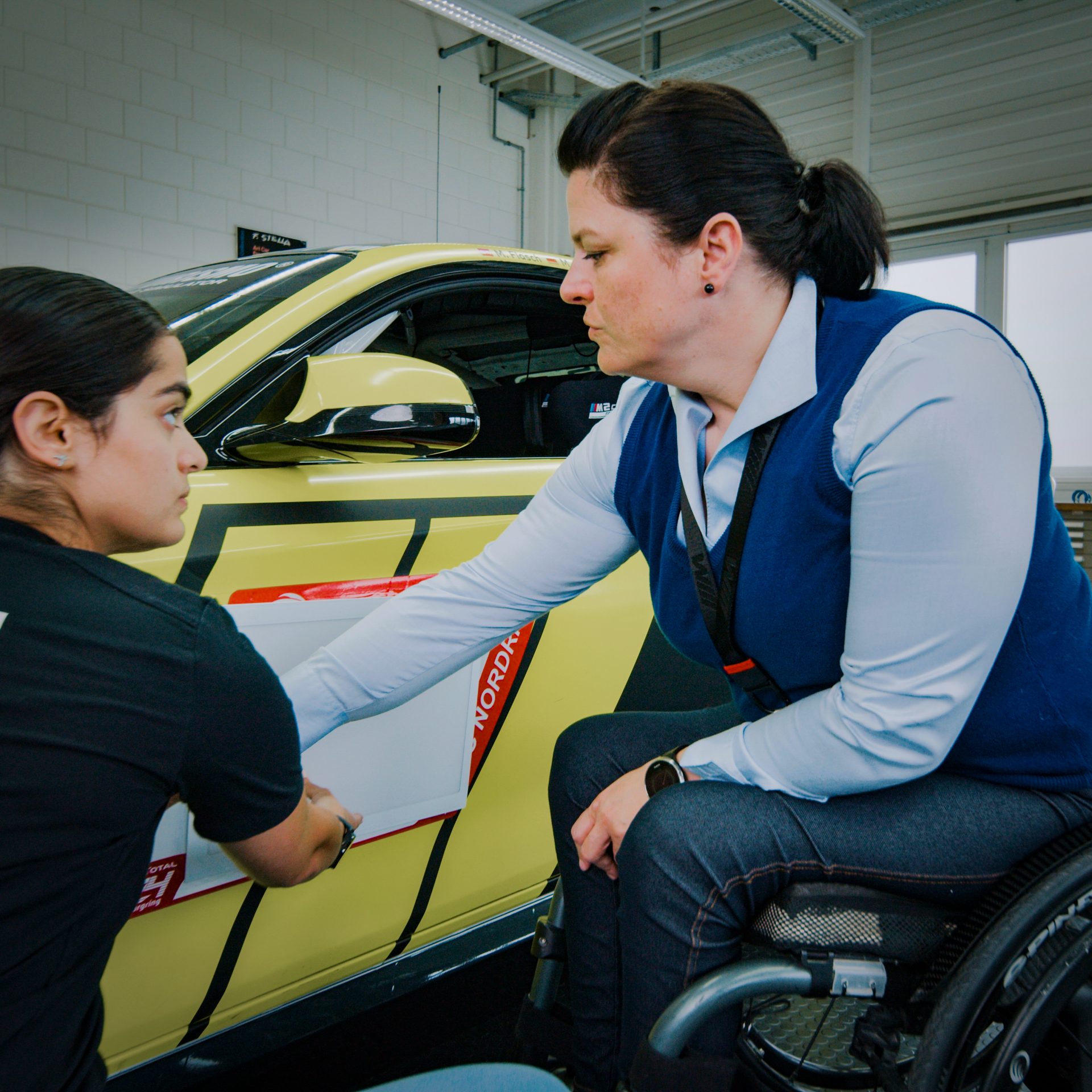 The female Head of Project Management for the Product Line M sitting in a wheelchair having a conversation with a colleague around a test vehicle.