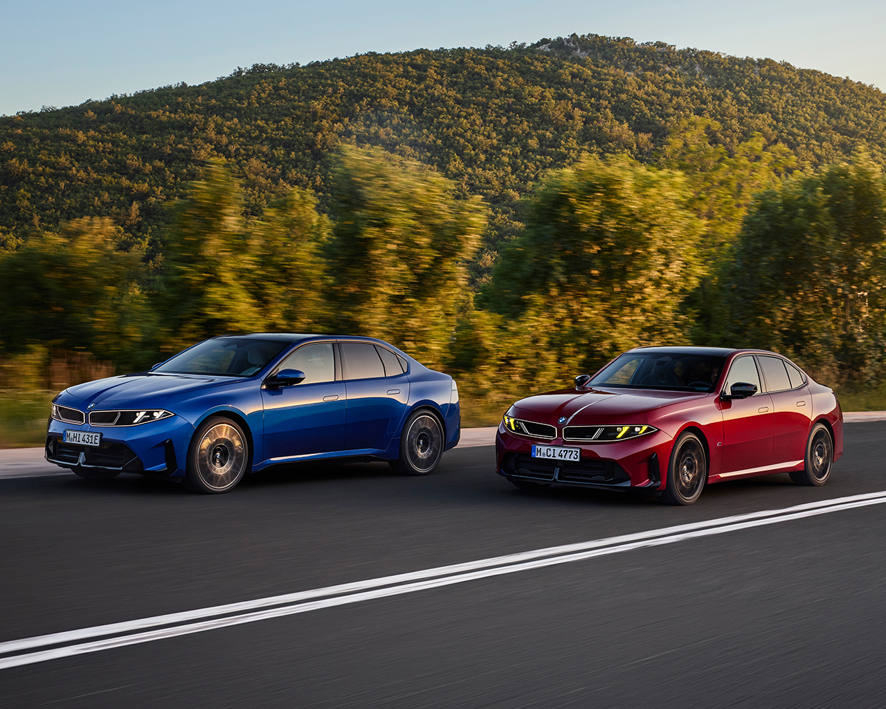 Ein blauer vollelektrischen BMW i3 50 xDrive (links) und ein roter BMW M350 xDrive fahren dynamisch und leicht versetzt nebeneinander auf einer Landstraße – aus schräger Frontperspektive aufgenommen; dahinter erhebt sich eine bewaldete Berglandschaft.