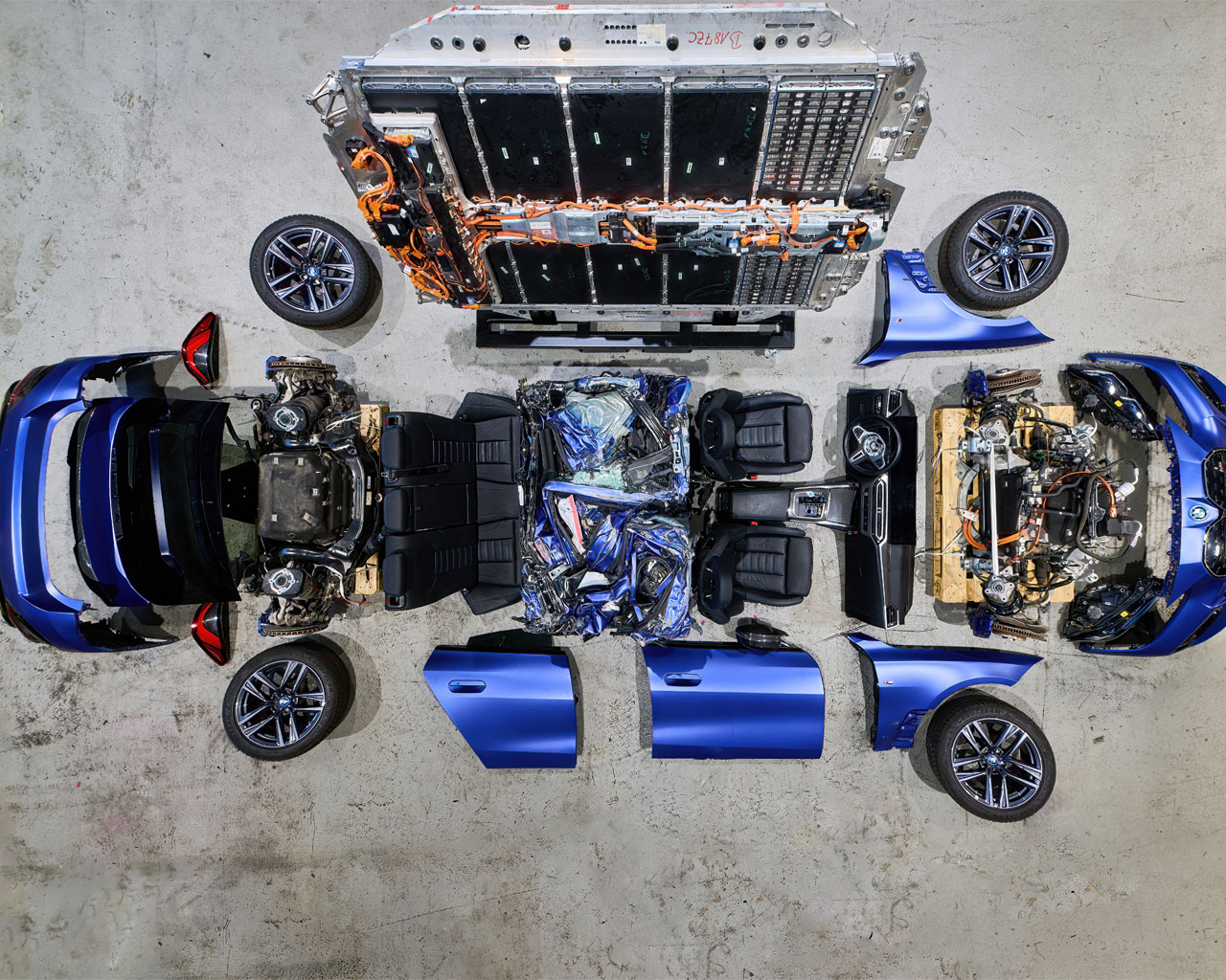 Disassembled blue car with body panels, seats, wheels, and engine components laid out on a concrete floor.