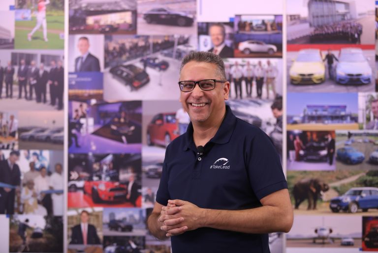 The picture shows a BMW Group employee standing in front of a picture wall.