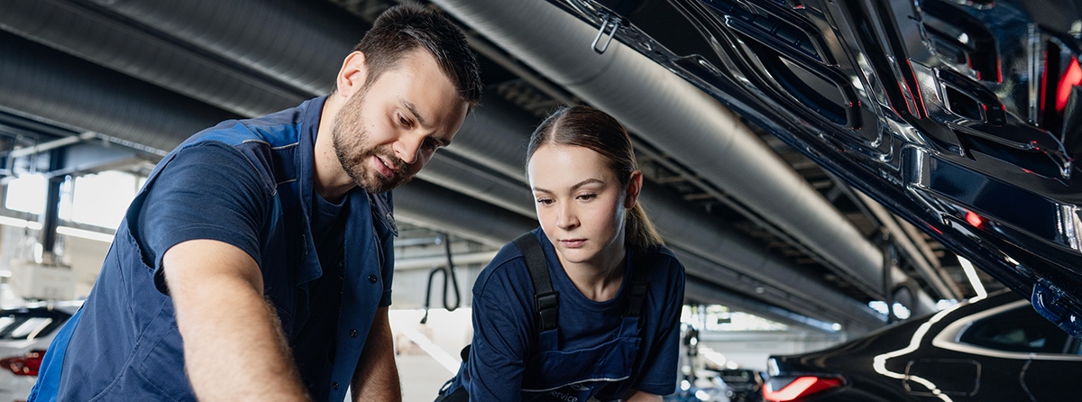 Ein Ausbilder und eine Auszubildende inspizieren den Motor eines Fahrzeugs mit offener Motorhaube.