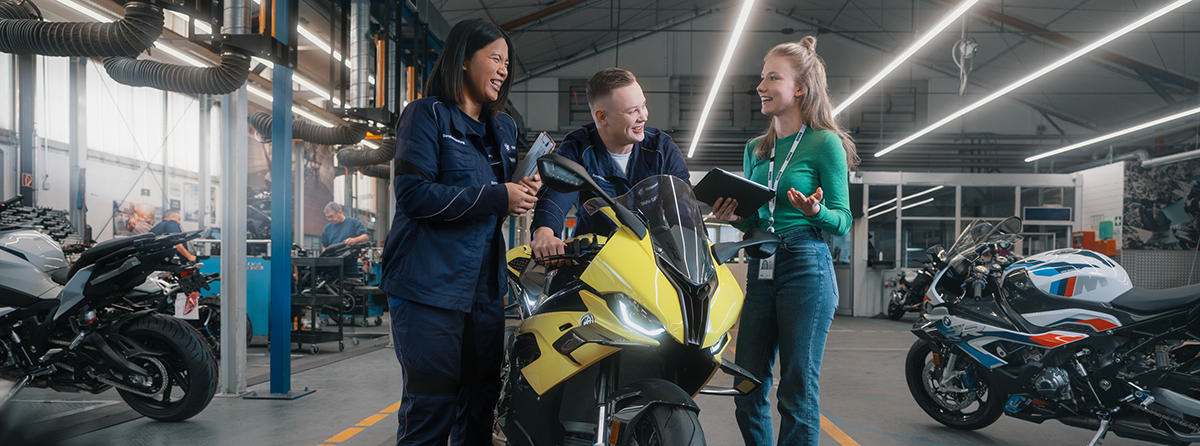 Drei Auszubildende stehen neben einem Motorrad und lachen.