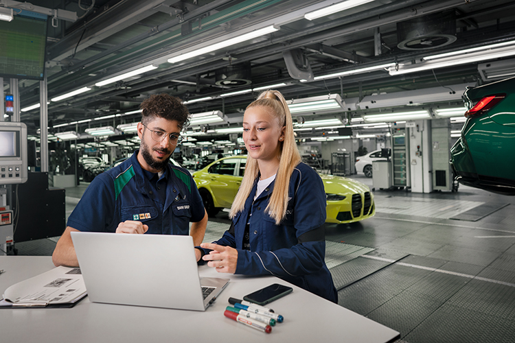 Zwei Auszubildende Mechaniker in einer Werkstatt besprechen Informationen am Laptop.