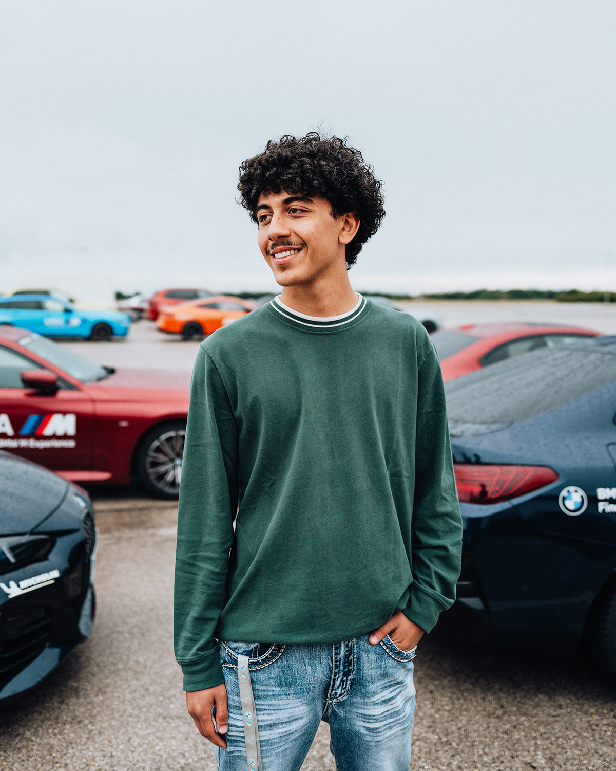 BMW Azubi Yahya vor verschiedenen BMW Fahrzeugen auf einer Teststrecke