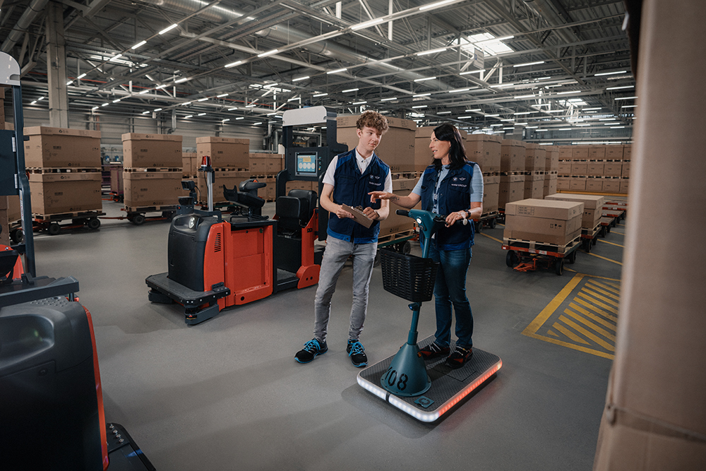 Zwei Auszubildende für Lagerlogistik sprechen in einem großen Lager mit Kartons bei der BMW Group.