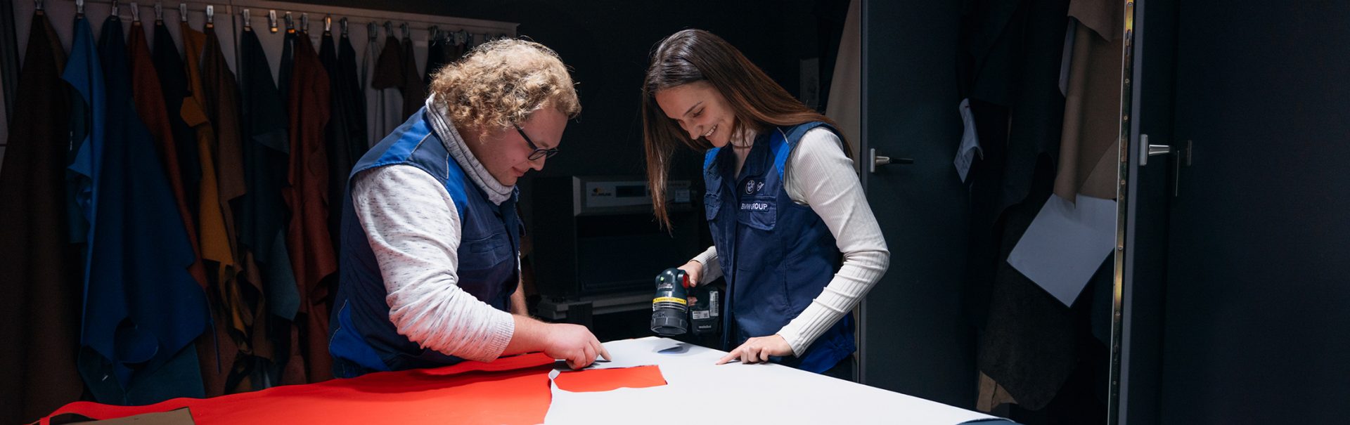 Eine Auszubildende und ein Experte im Bereich Fahrzeuginterieur arbeiten an einem Tisch mit Leder und Werkzeugen bei der BMW Group.