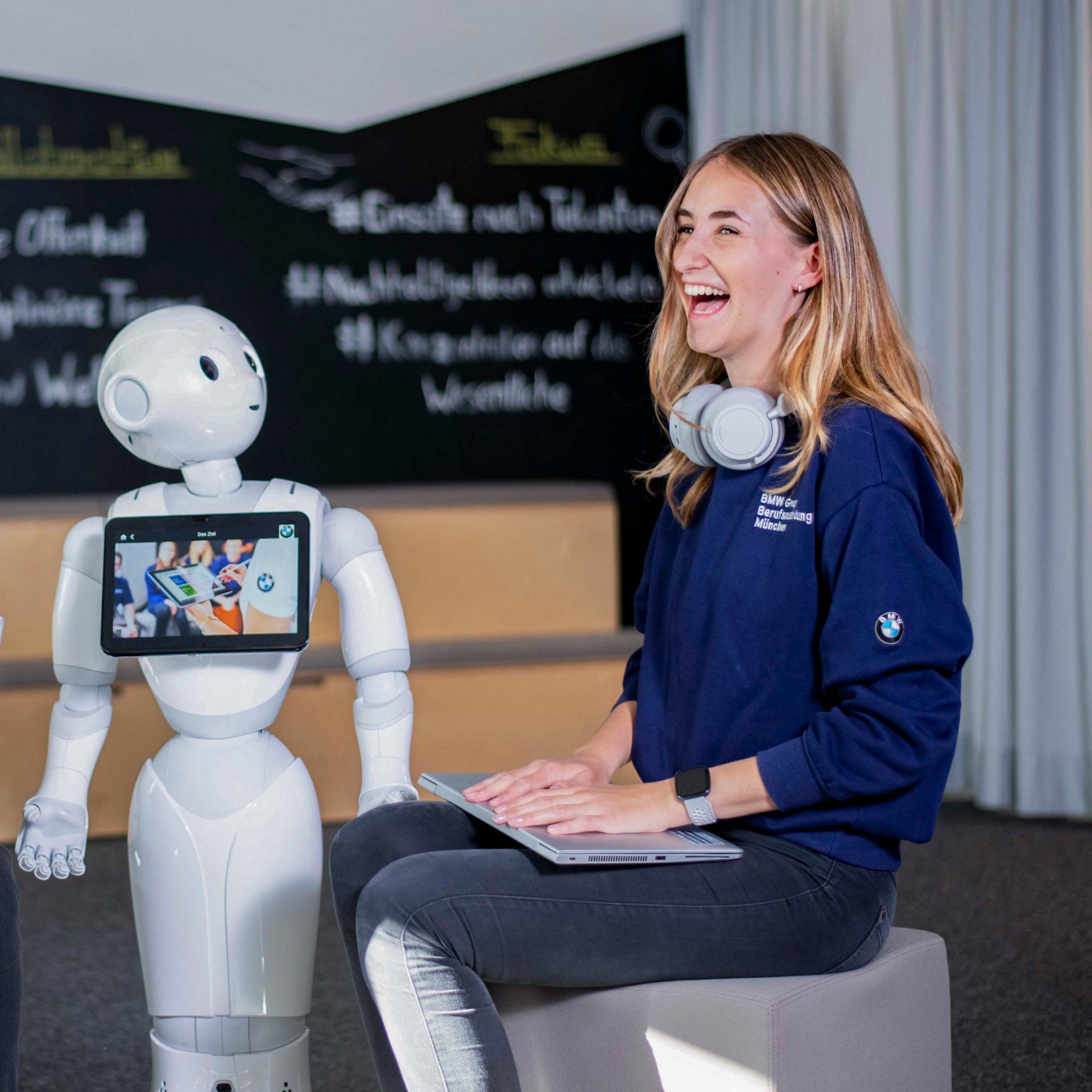 Eine junge Auszubildende bei der BMW Group sitzt neben einem Roboter und lächelt.