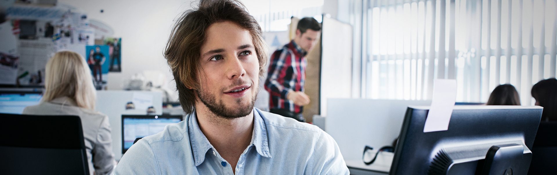 Nachdenklicher Auszubildender als E-Commerce Kaufmann bei der BMW Group in einem Büro mit Computern im Hintergrund.