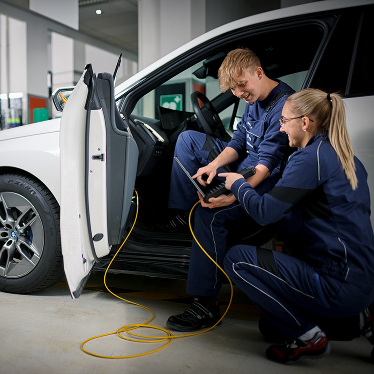 Zwei auszubildende Mechatroniker bei der BMW Group arbeiten an einem Auto mit einem Laptop.