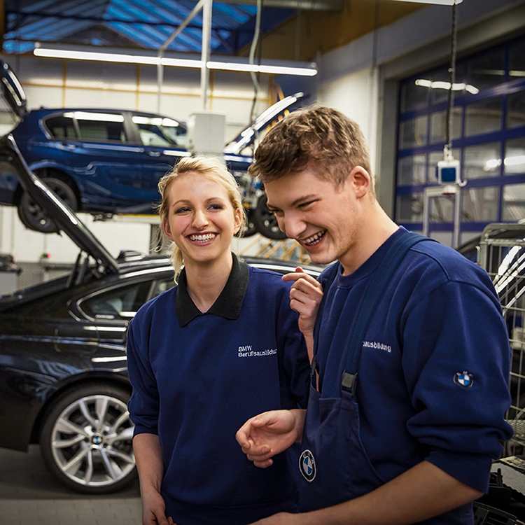  Junge Auszubildende bei der BMW Group in blauen Overalls in einer Autowerkstatt mit BMW-Fahrzeugen im Hintergrund.