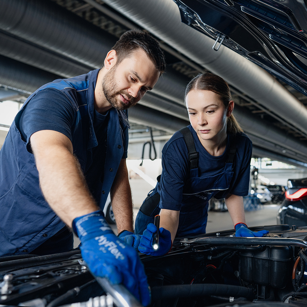  Junge Auszubildende bei der BMW Group in blauen Overalls in einer Autowerkstatt mit BMW-Fahrzeugen im Hintergrund.