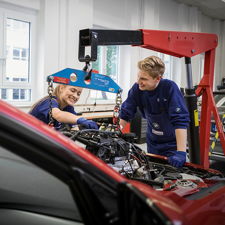 Zwei Mechatroniker arbeiten in einer Werkstatt der BMW Group im Motorraum eines Auto.