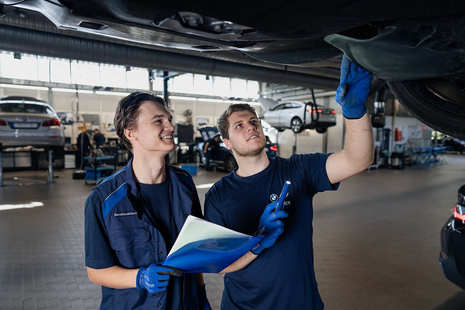 Zwei Auszubildende in der BMW Werkstatt lachen und arbeiten zusammen.