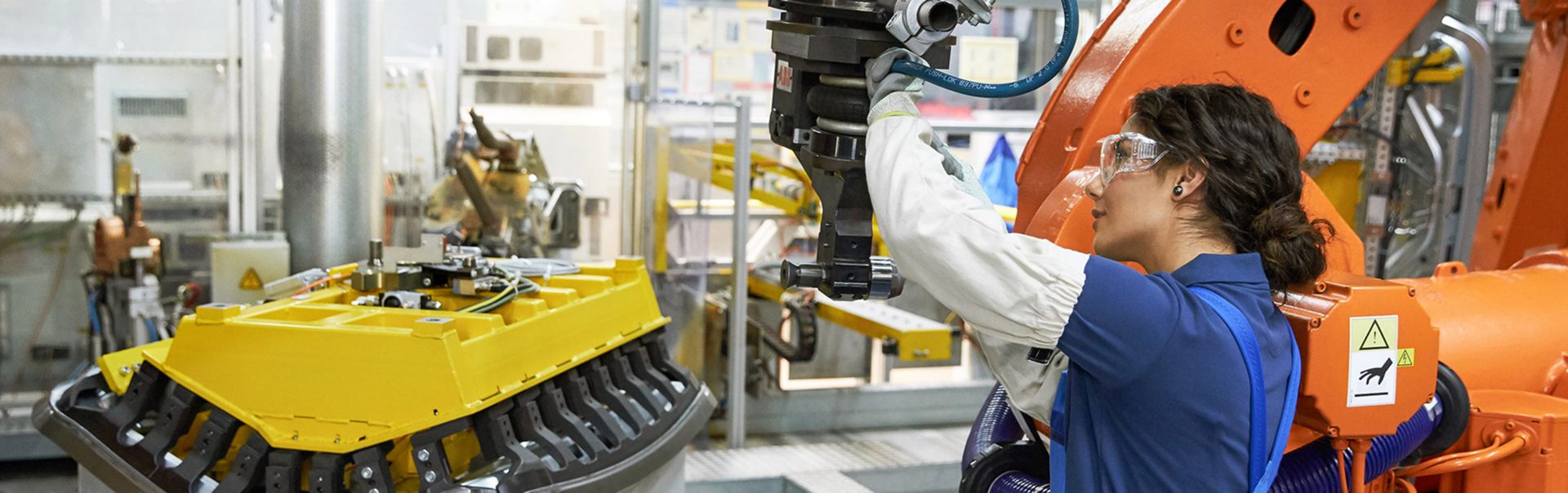 Person in blauer Arbeitskleidung bedient einen orangen Industrieroboter in einer Werkhalle mit gelbem Maschinenteil.