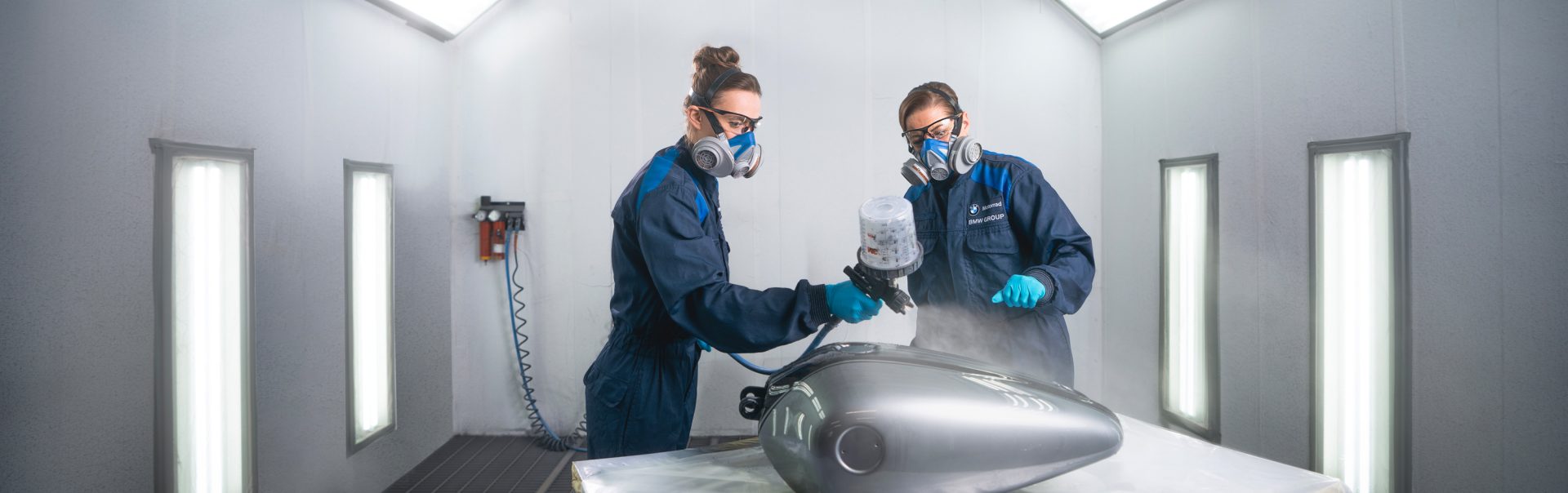 Zwei Verfahrensmechaniker für Beschichtungstechnik bei der BMW Group lackieren ein Motorradteil in einer professionellen Lackierkabine.