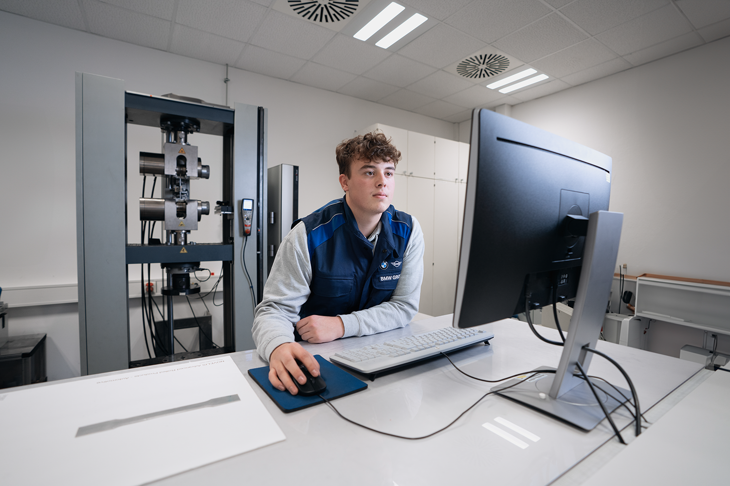 Auszubildender arbeitet am PC neben einer Werkstoffprüfanlage.