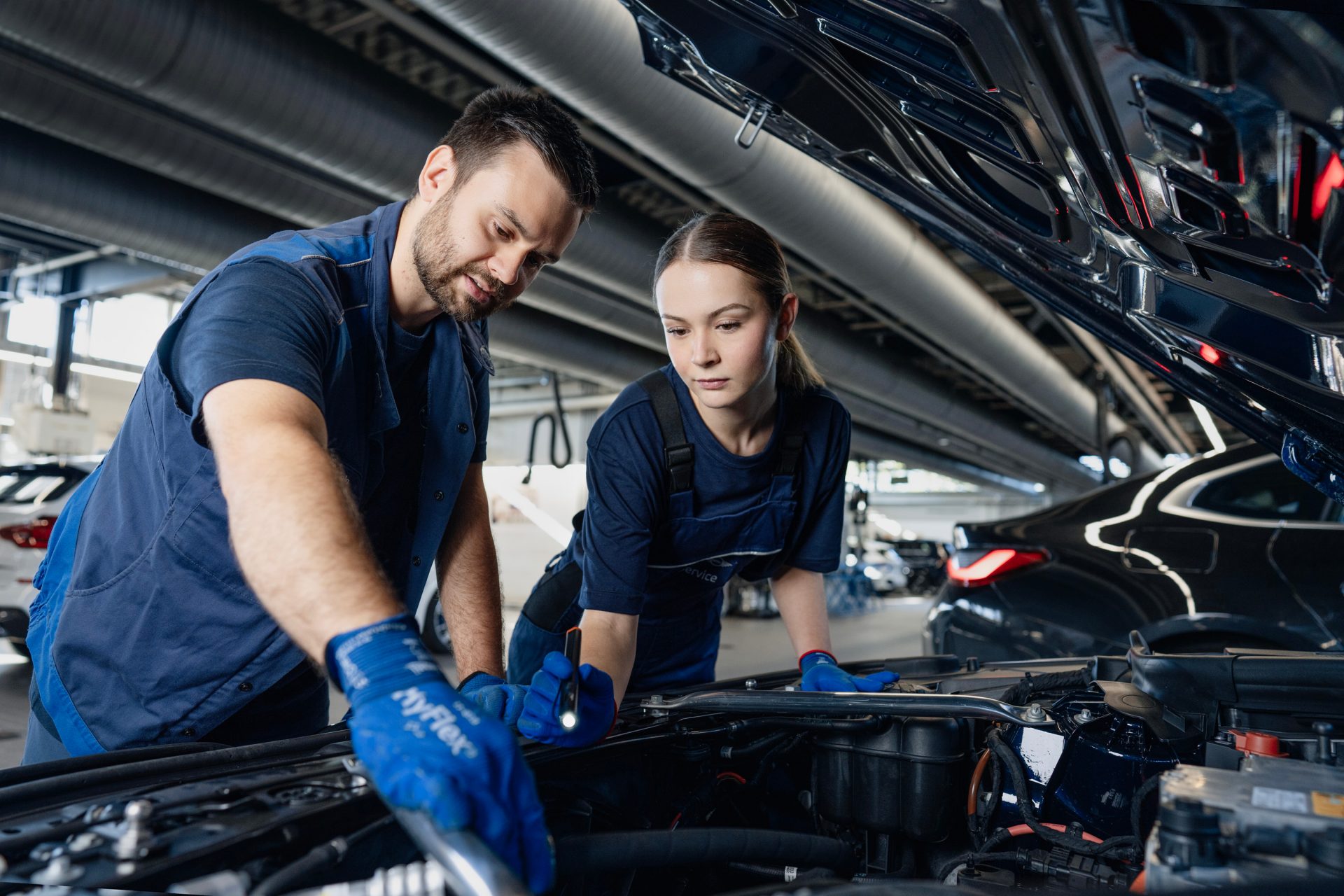 Zwei Auszubildende in einer BMW-Werkstatt, die an einem Laptop arbeiten. Im Hintergrund ist ein BMW iX zusehen, an dem zwei weitere Auszubildende arbeiten.