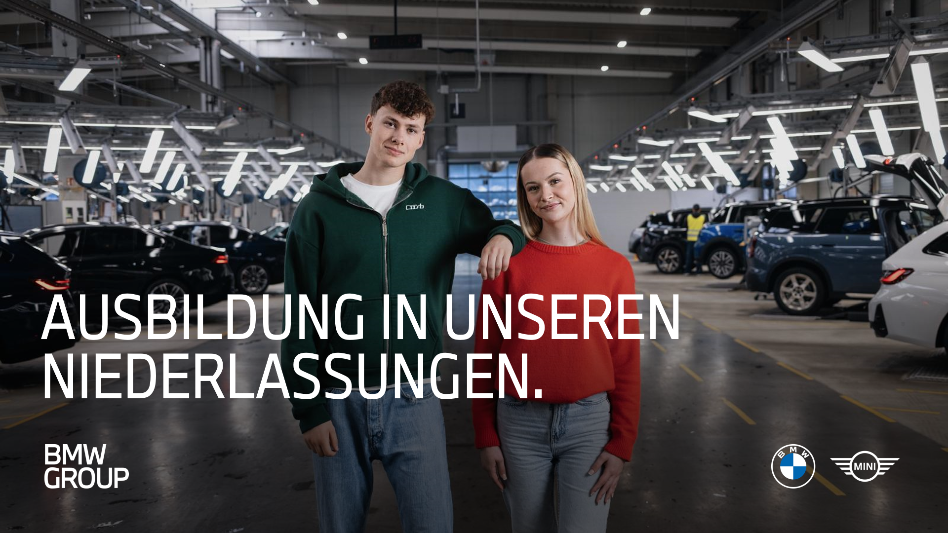 Zwei Azubis stehen lässig in einer Kfz-Werkstatt und lachen in die Kamera