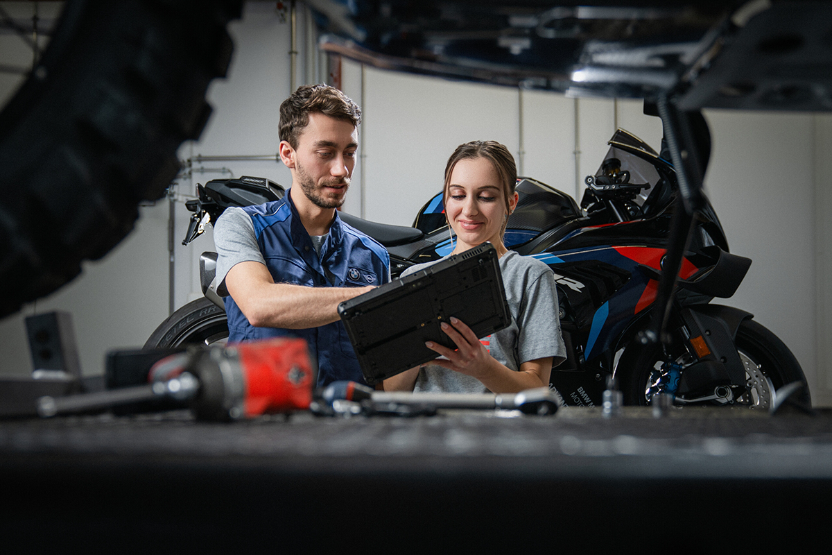 Ein Ausbilder erklärt einer Schülerpraktikantin etwas auf einem Tablet, Motorrad im Hintergrund