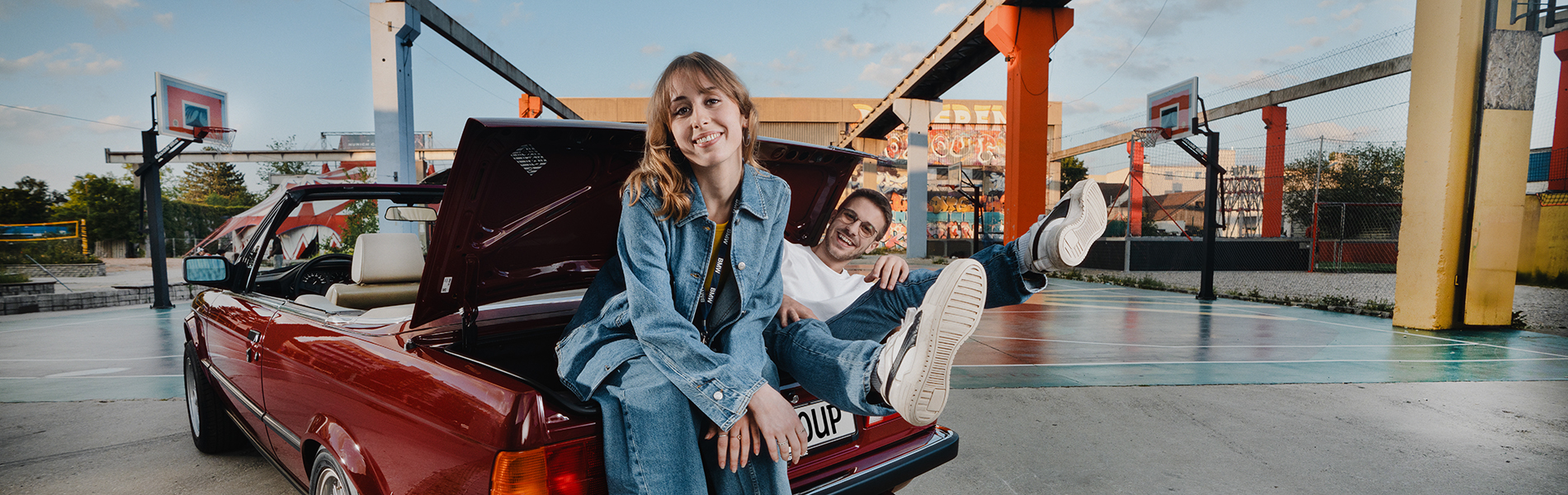 Zwei Azubis sitzen im Kofferraum eines BMW Oldtimers und lachen herzlich