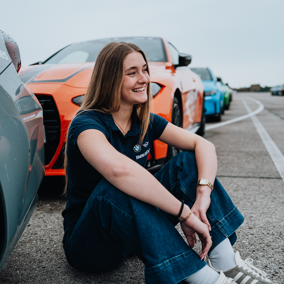 Duale Studentin Ludovica sitzt lachend am Boden vor einem orangen BMW M3