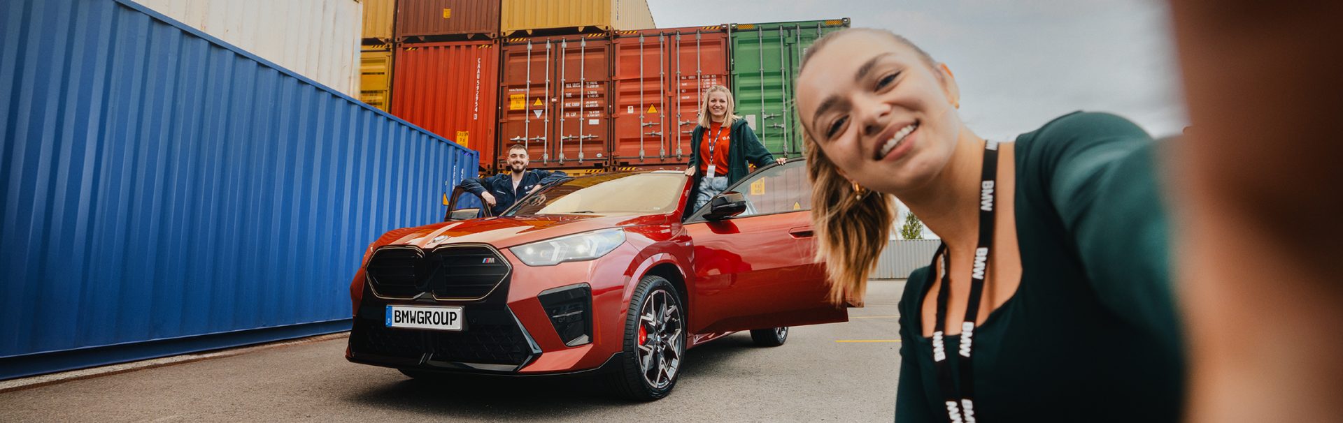 Drei duale Studenten der BMW Group posieren mit einem BMW X2 vor bunten Containern.