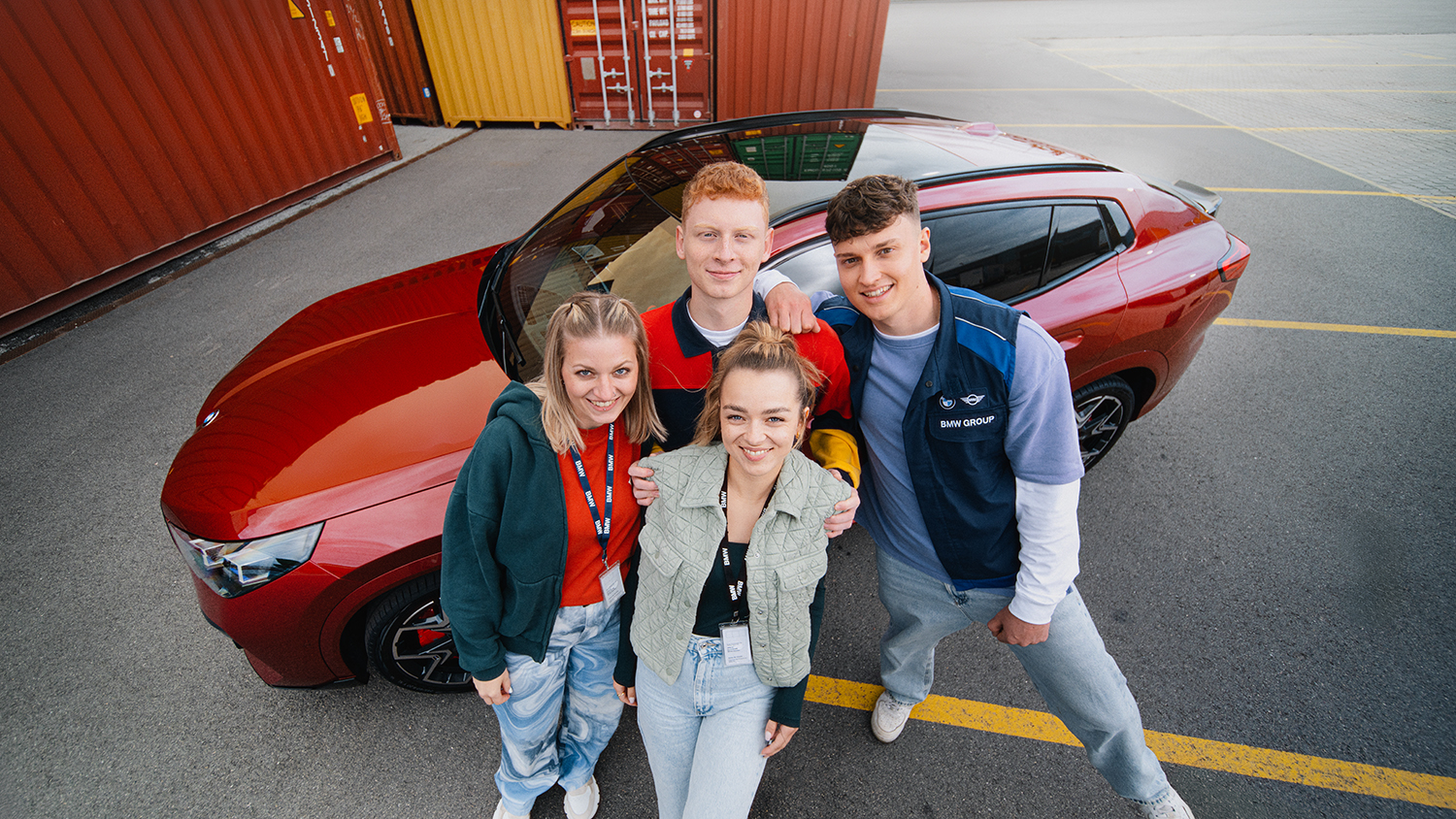 Drei duale Studenten der BMW Group posieren mit einem roten BMW X2 vor bunten Containern.