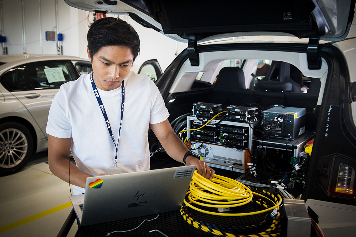 Azubi bei der Arbeit an einem Auto, in dem im Kofferraum viel Technik verbaut ist