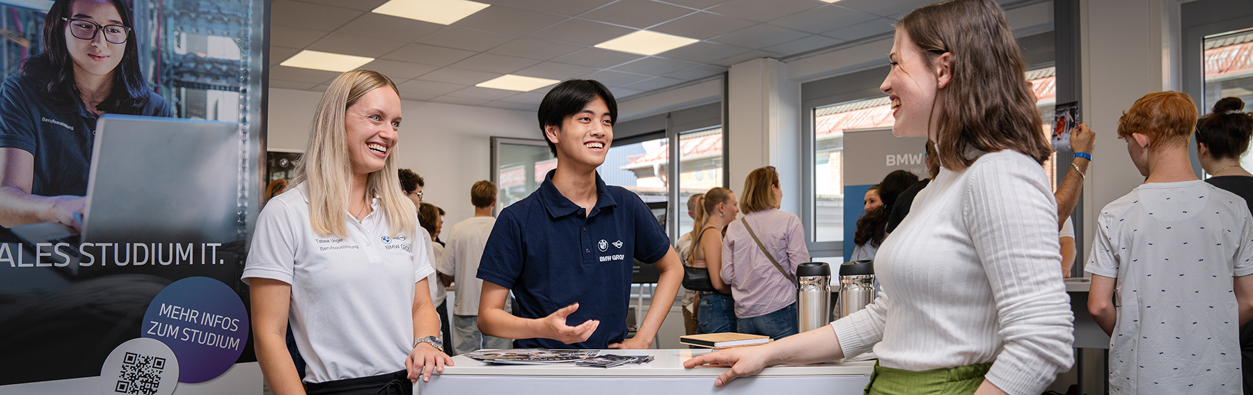 Eine Interessentin an einem Messestand beim Berufsinfotag unterhält sich mit einem Azubi und seiner Ausbilderin