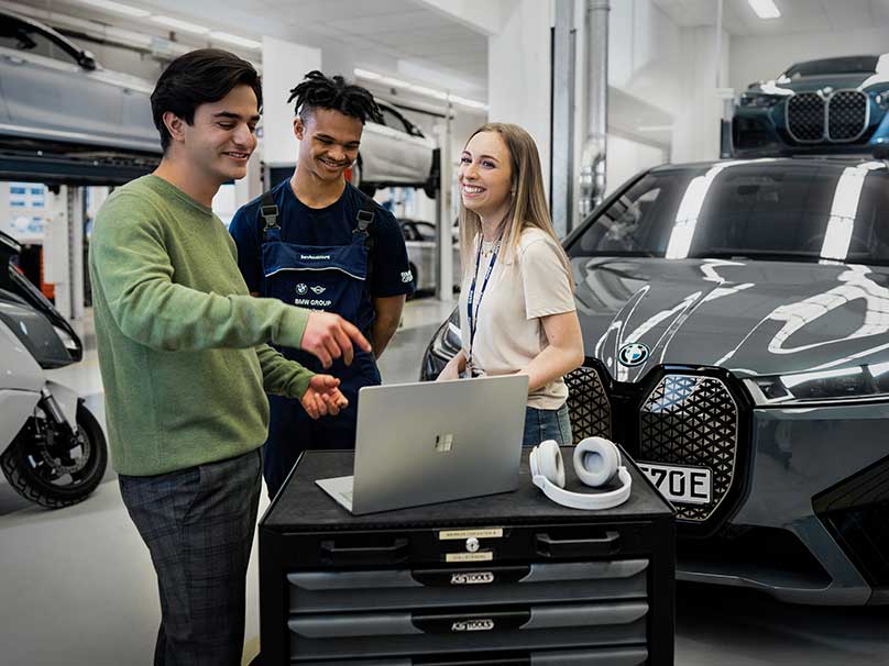 Das Bild zeigt drei lachende Auszubildende in einer BMW Werkstatt. 