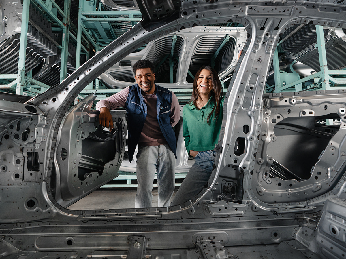 Zwei Auszubildende der BMW Group stehen in der Produktionshalle und lächeln durch eine Fahrzeugkarosserie hindurch.