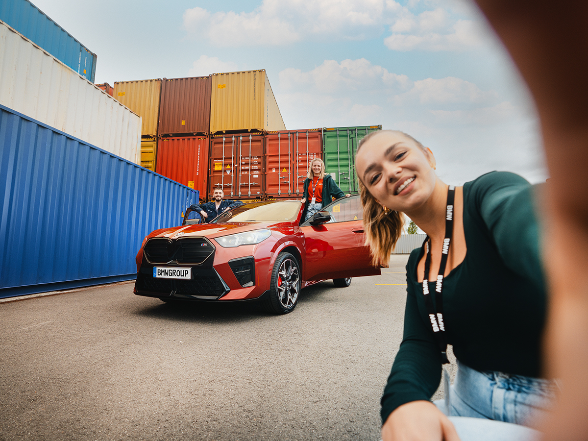 Drei duale Studenten der BMW Group posieren mit einem roten BMW X2 vor bunten Containern.
