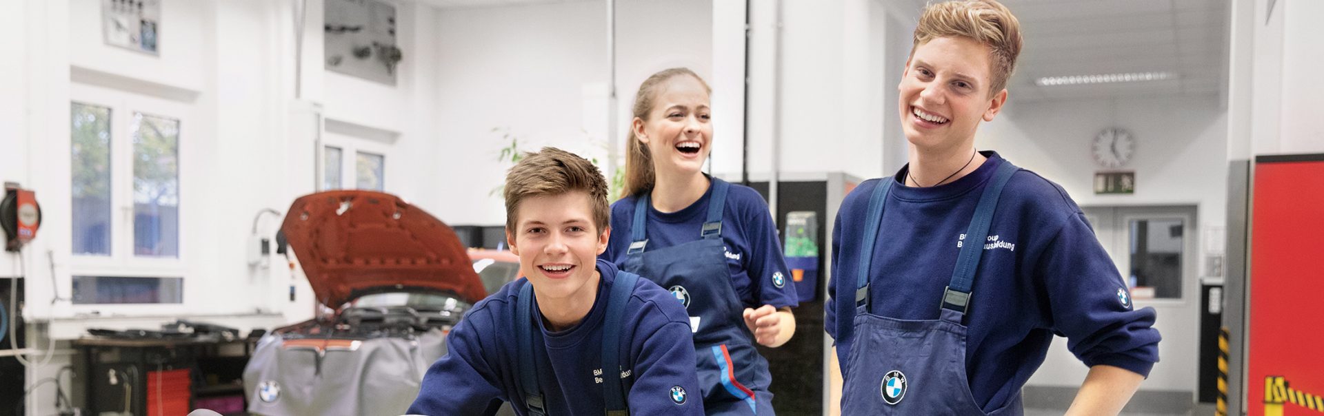 Drei Schüler stehen zusammen in BMW Arbeitsklamotten in einer Werkstatt, einer von ihnen sitzt auf einem Motorrad.