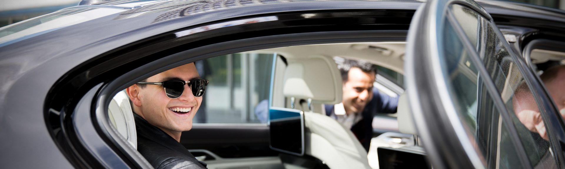 Ein junger Mann sitzt auf der Rückbank eines BMW Fahrzeuges, zwei weitere Männer schauen jeweils durch Fahrer- und Beifahrertüre in das Fahrzeug hinein.