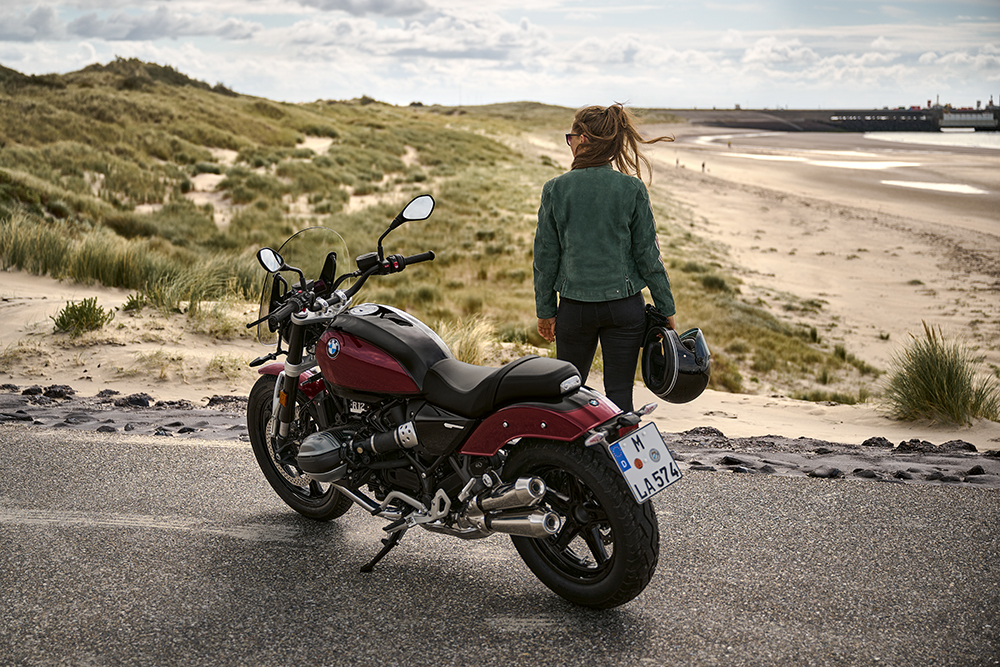 Eine Frau steht neben ihrem Motorrad an einem Sandstrand.