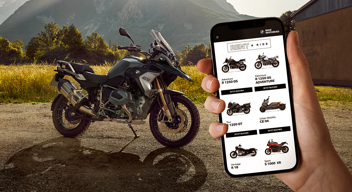 Eine Hand die ein Smartphone hält, auf welchem verschiedene Motorradmodelle angezeigt werden. Im Hintergrund steht ein BMW Motorrad.
