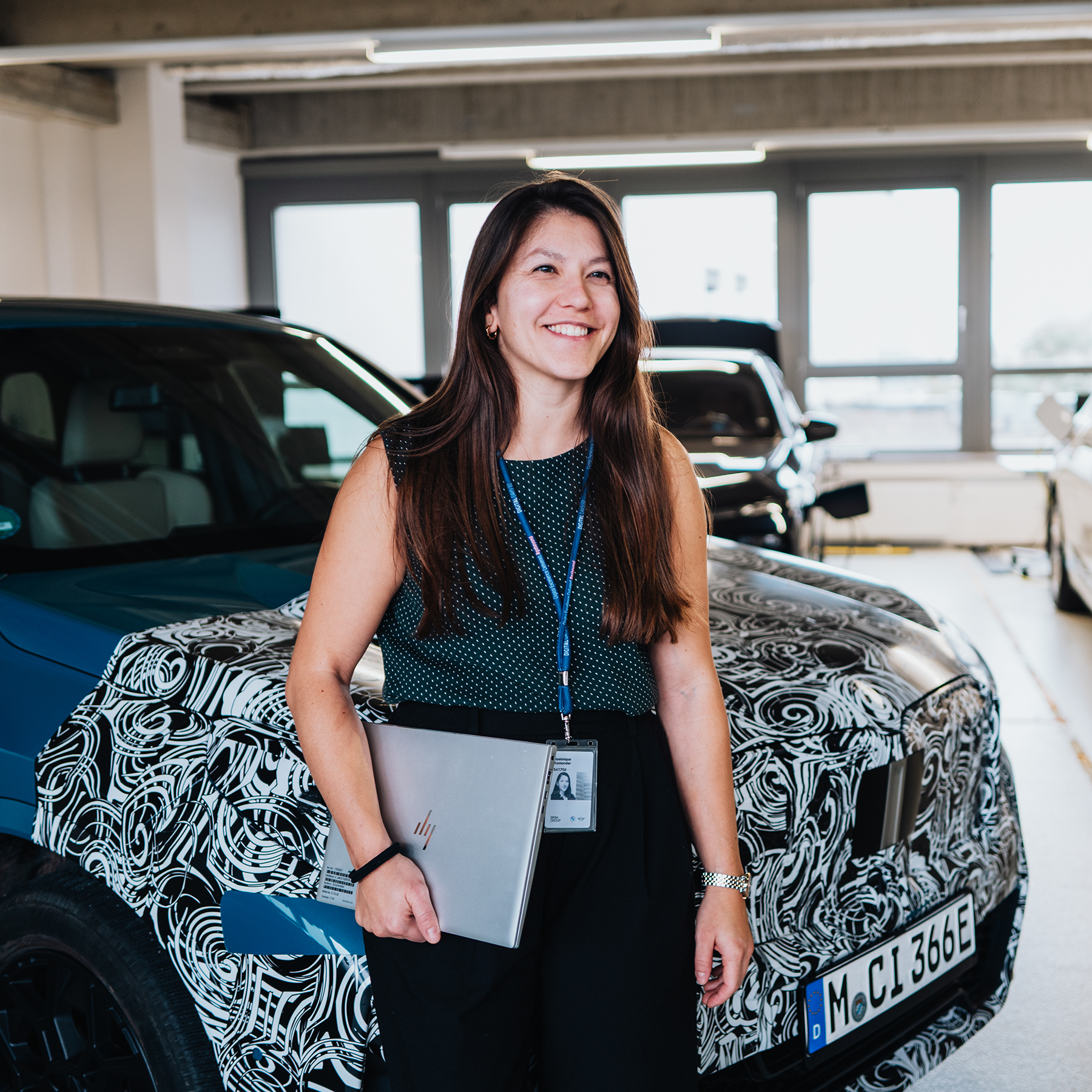 Dominique lehnt sich an einen BMW IX2 und lächelt in die Kamera.