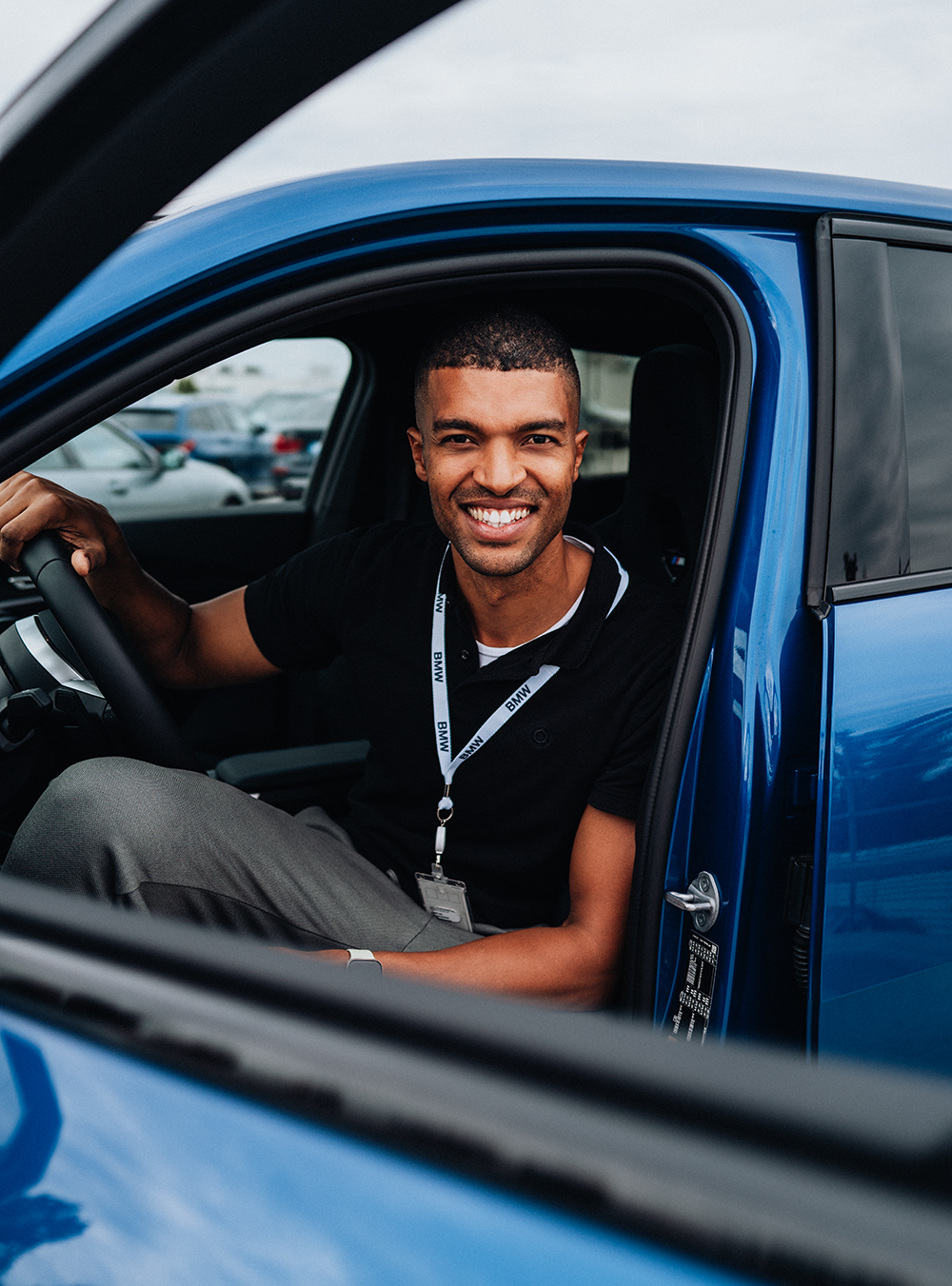 Ingenieur der BMW Group sitzt auf dem Fahrersitz eines blauen BMW iX2 und lächelt auf dem Fenster in die Kamera.