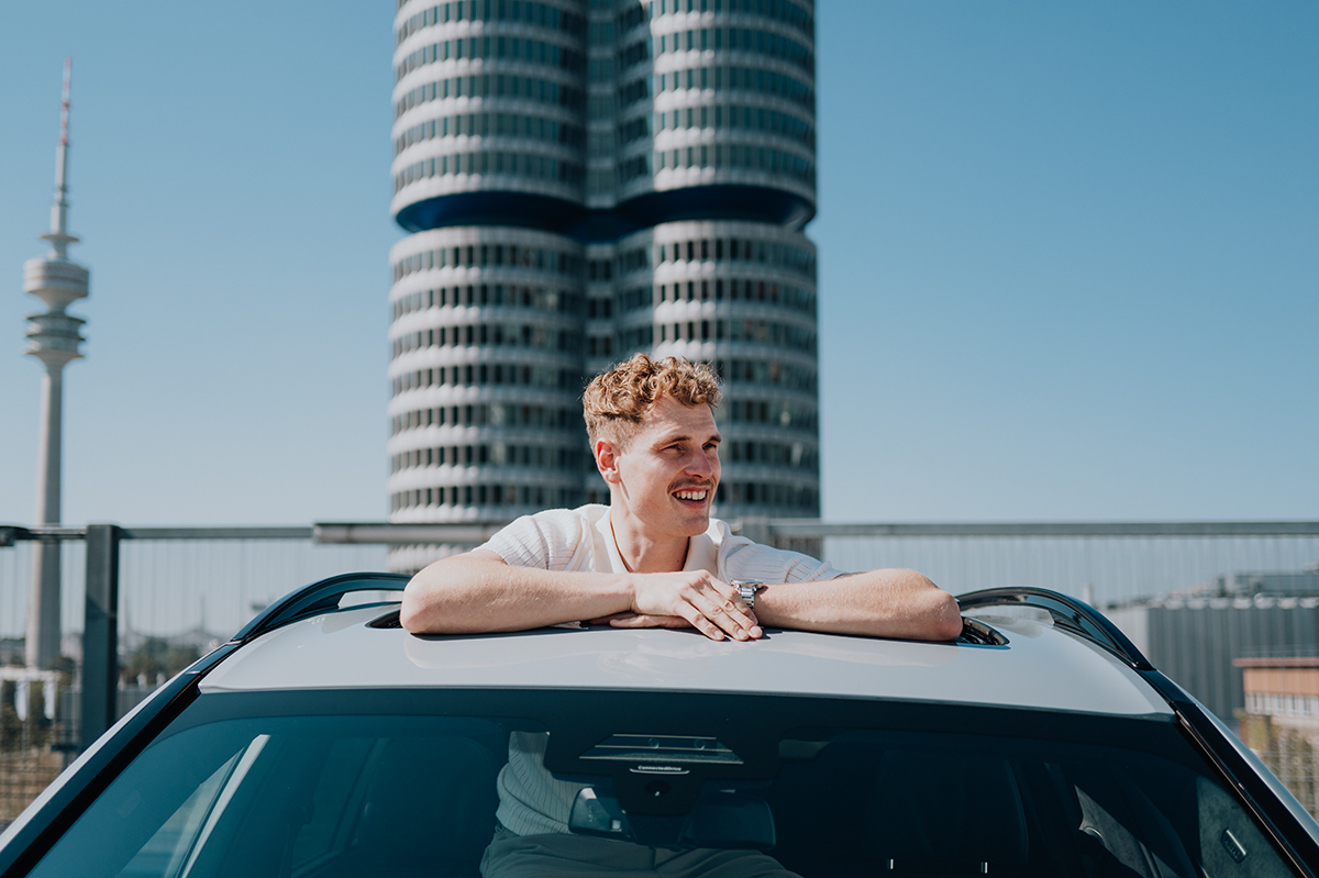 Ingenieur René schaut aus dem Schiebedach des BMW X7 mit BMW Vieryzlinder Gebäude im Hintergrund