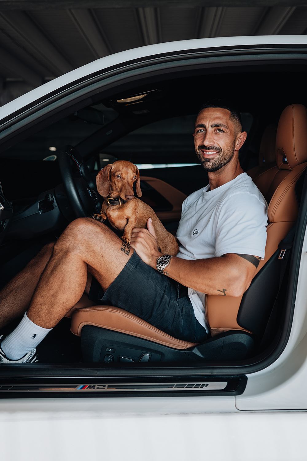 BMW Mitarbeiter mit seinem braunen Dackel auf dem Fahrersitz eines weißen BMW M2.