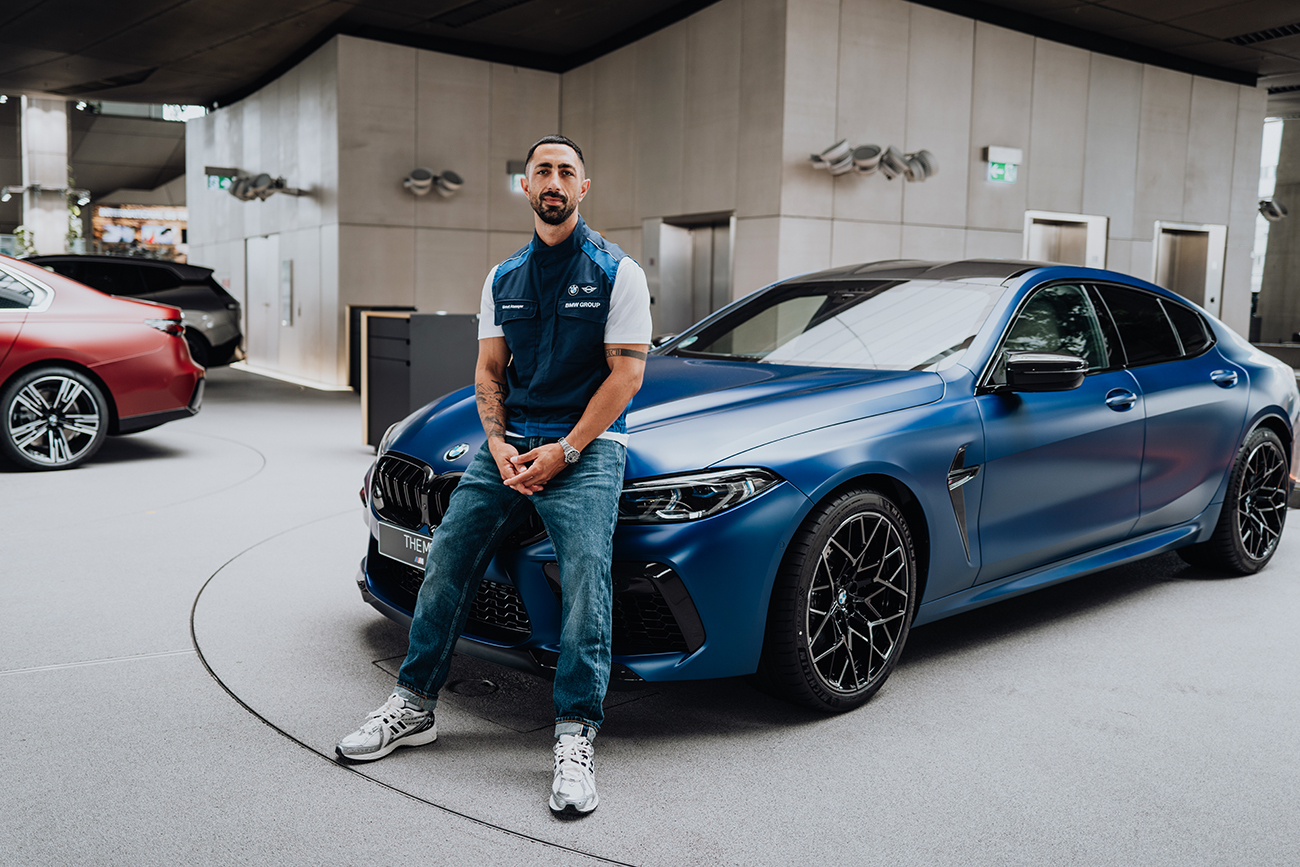 BMW Mitarbeiter lehnt an einem Matt-blauen BMW M8 in der BMW Welt.