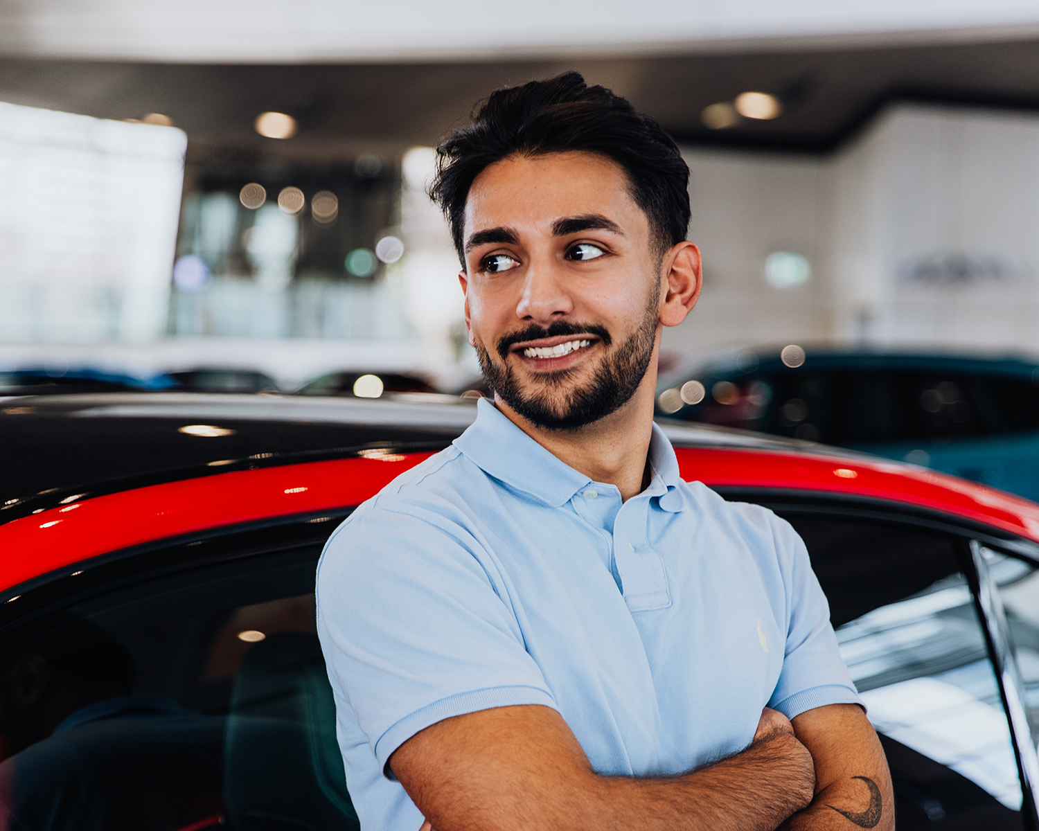 Yasar lehnt mit verschränkten Armen an einen roten BMW M5 und schaut in die Ferne.