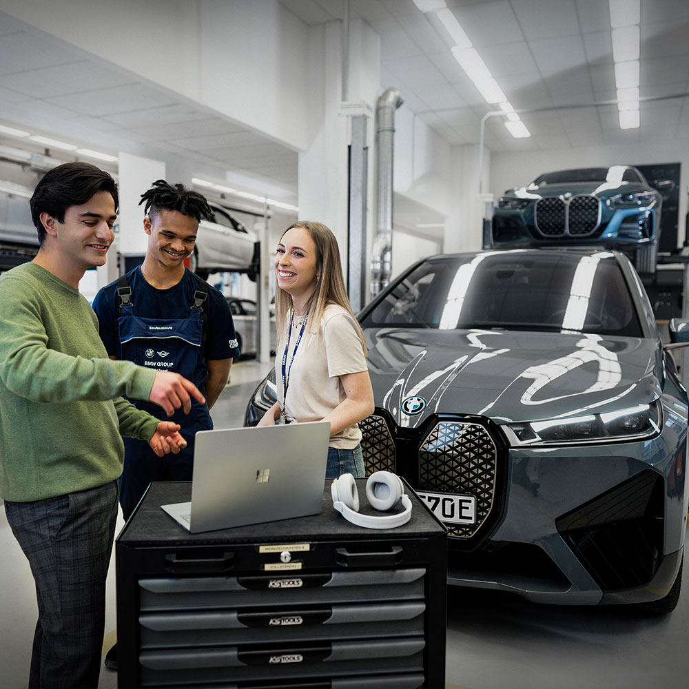 The picture shows three apprentices pointing at a laptop while standing in front of a vehicle.