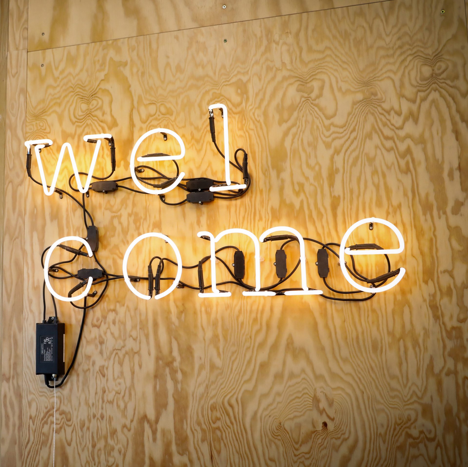 The picture shows the word "Welcome" in neon letters on a wooden wall.