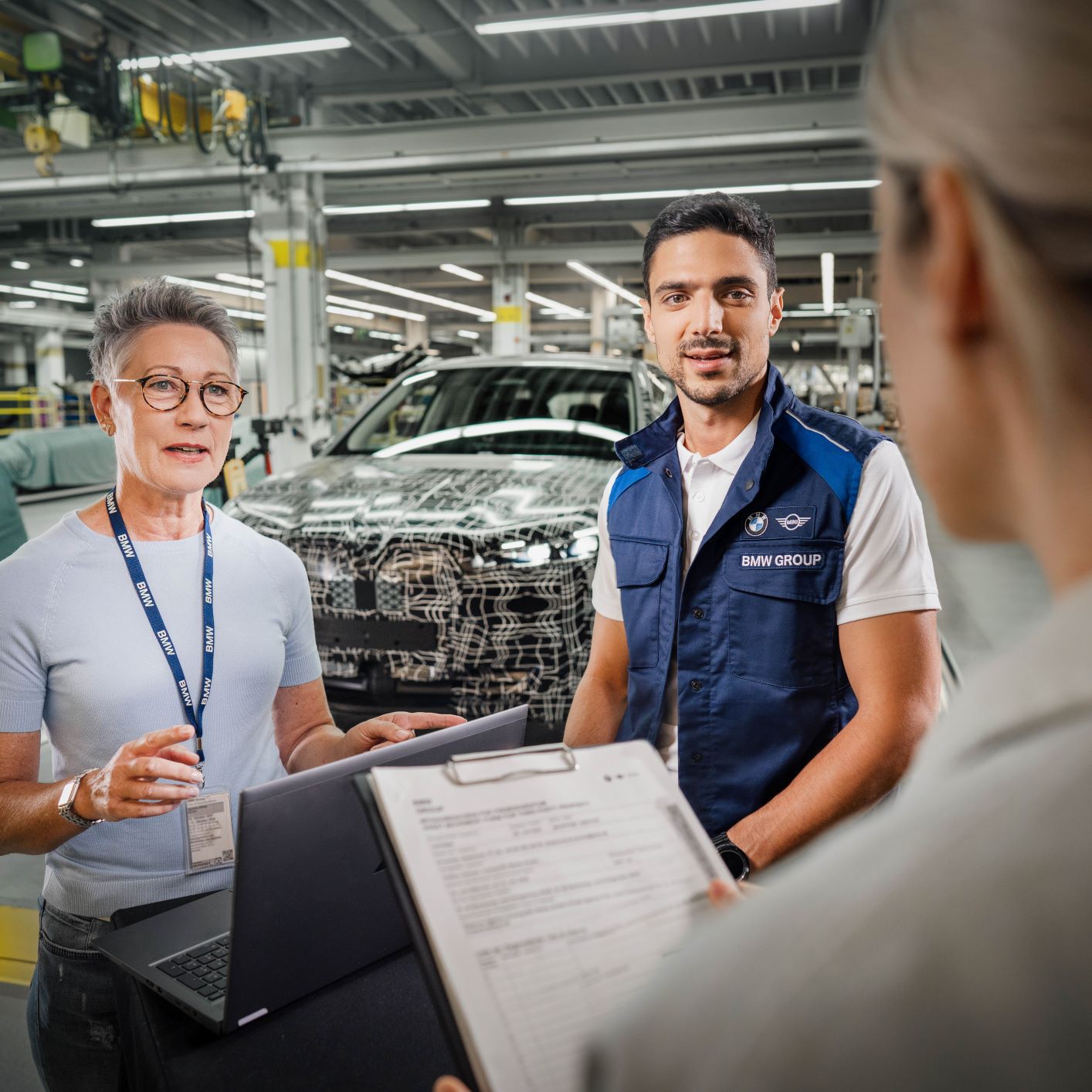 Three BMW engineers in a factory setting with a BMW vehicle in the background..