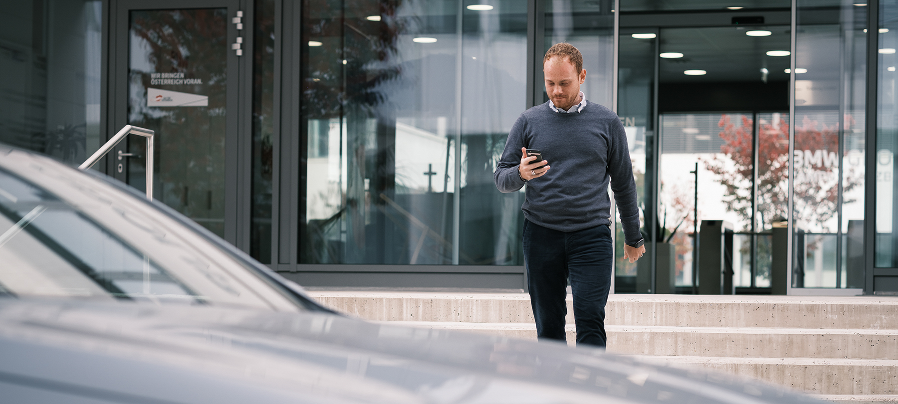 A employee leaving the BMW Group office builing on his way to his car looking at his smartphone.